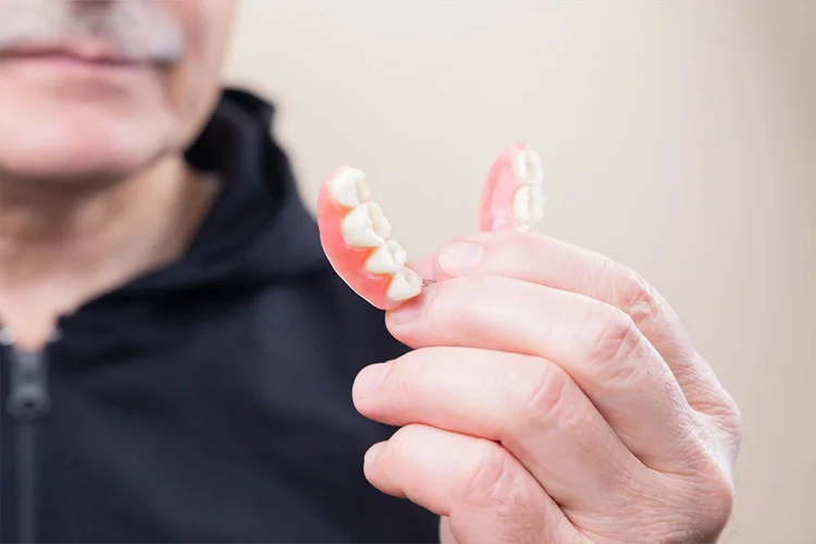 A person holds a set of dentures, showcasing their detail, with a neutral background.