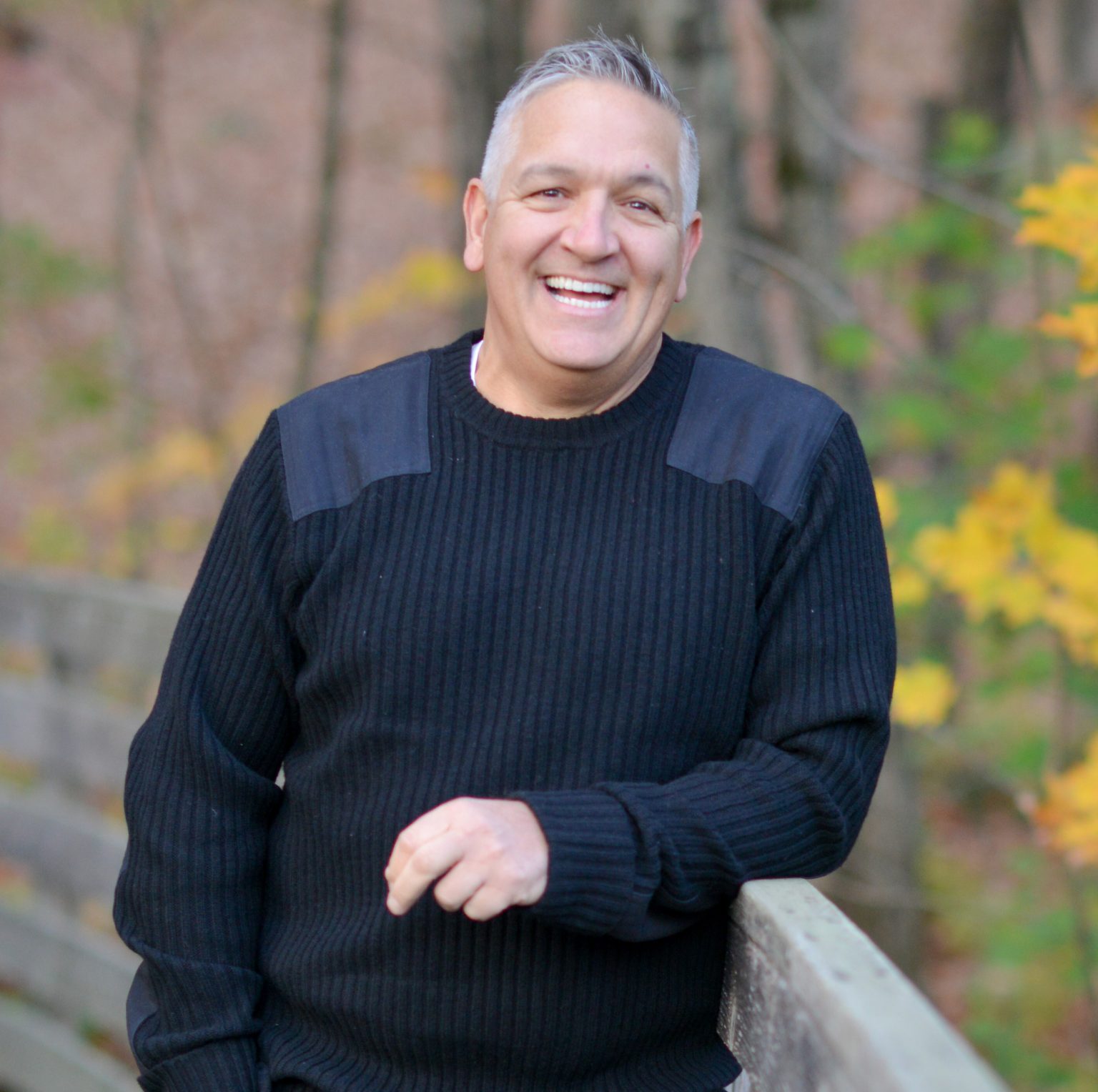 Smiling man in a black sweater stands by a wooden railing, surrounded by autumn foliage in a peaceful setting.