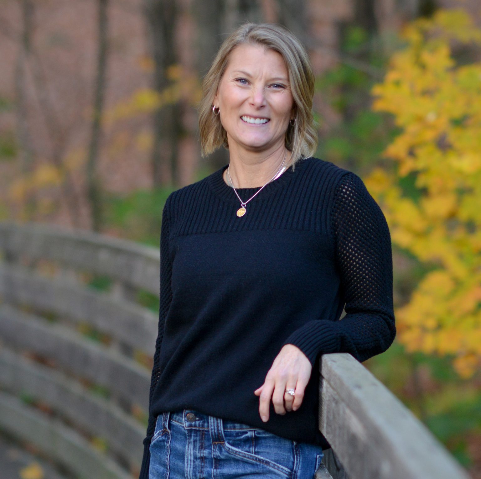 A smiling woman in a black sweater leans against a wooden railing, surrounded by autumn foliage.
