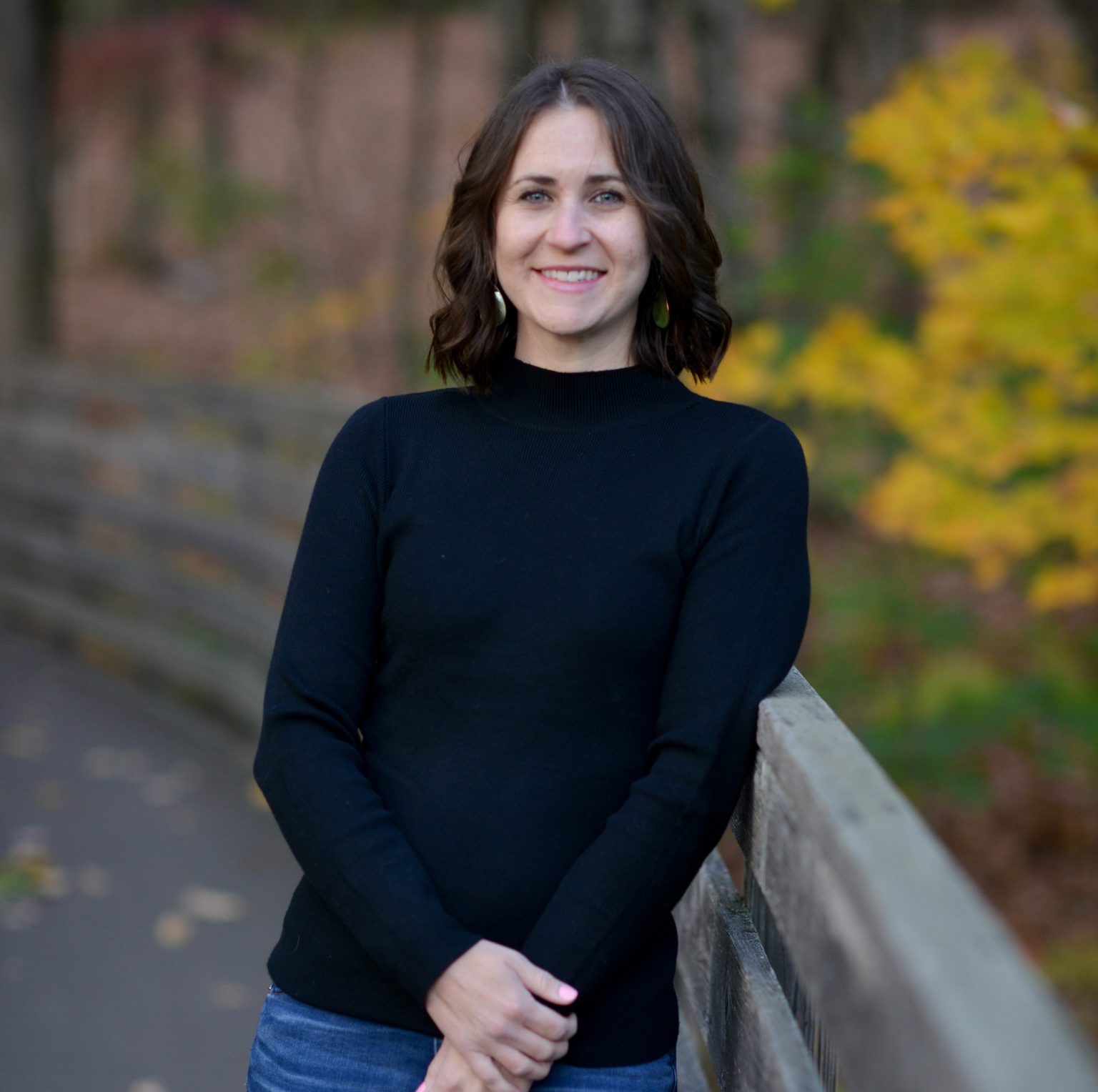 Smiling person with shoulder-length dark hair, wearing a black shirt, standing by a wooden railing in a fall setting.