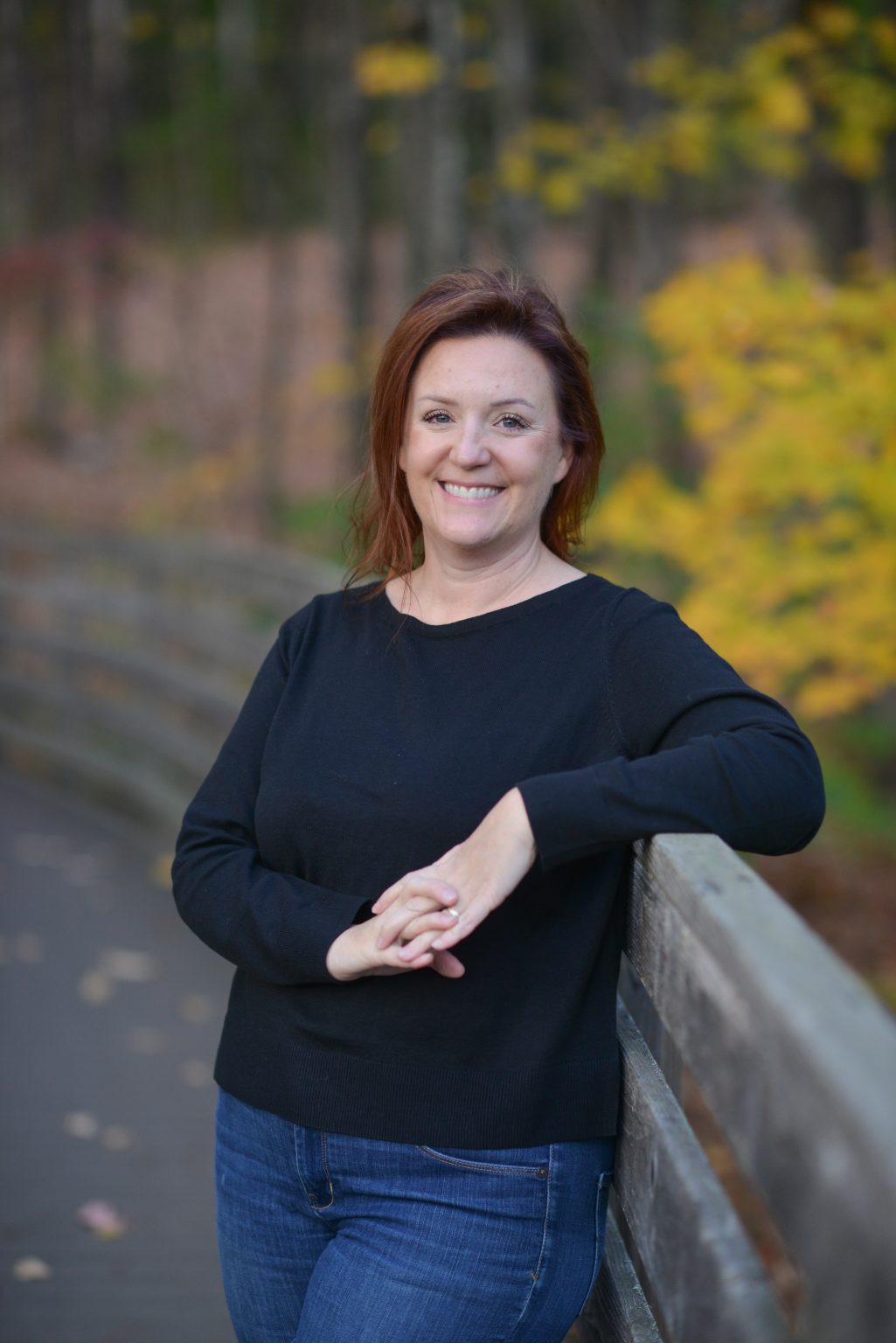 A woman in a black sweater leans on a wooden railing, smiling amidst a colorful autumn backdrop.
