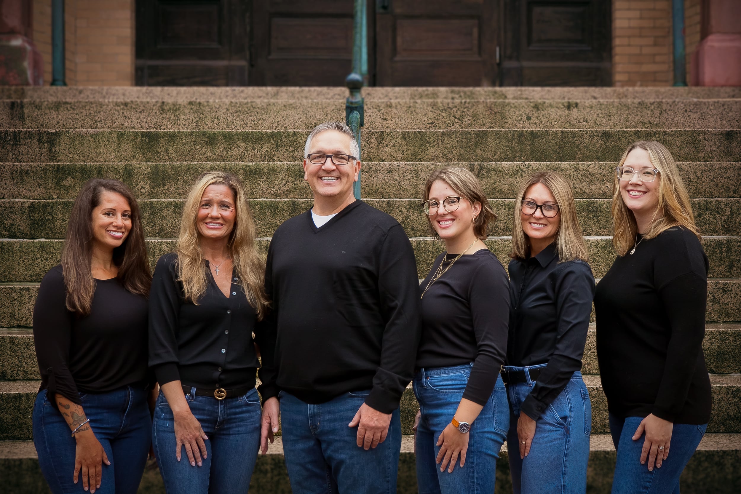 A group of six people, dressed in black shirts and jeans, pose together on outdoor steps.