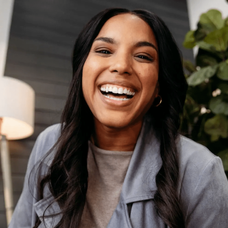 Smiling woman with long hair, wearing a grey jacket, seated in a cozy room with a lamp and plants in the background.