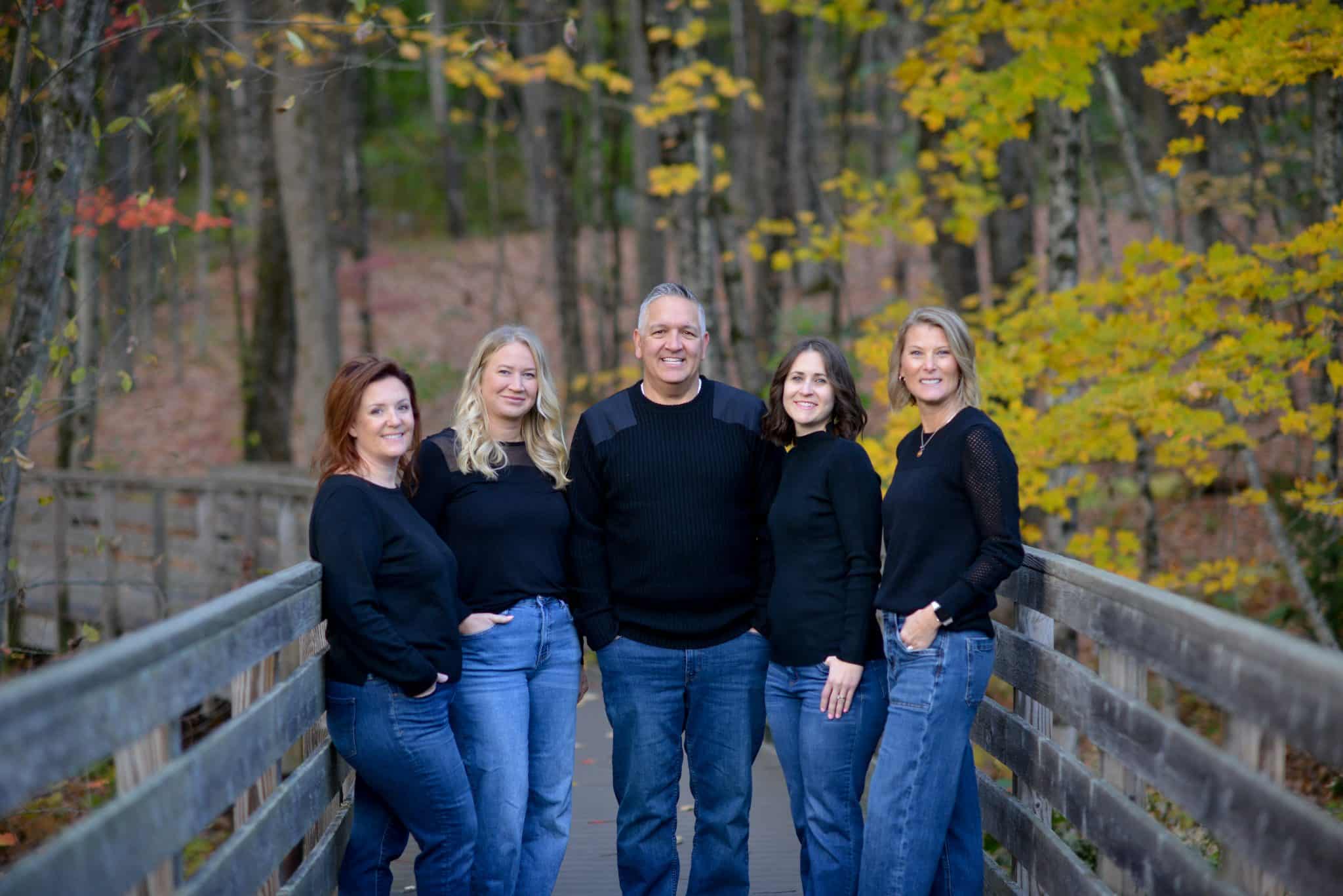 A group of five people wearing black sweaters stand together on a wooden bridge, surrounded by autumn foliage.